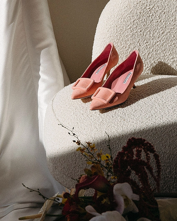 Bridal shoes in pink wedding heels with buckle detail resting on a textured armchair, framed by gown fabric and bouquet in window light