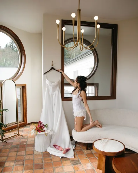 Wedding dress moment with bride in lingerie reaching for a gown on a hanger, pink heels and bouquet by arched windows in soft light