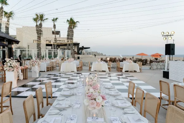 Wedding reception decor with black and white dance floor under string lights, long banquet tables set on a beachfront patio with palms and ocean view