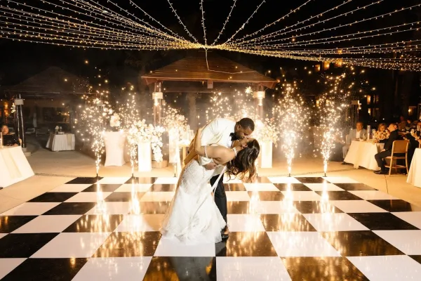 First dance as groom dips bride for a kiss on a black and white floor, cold spark fountains and string lights glowing on an outdoor patio at night
