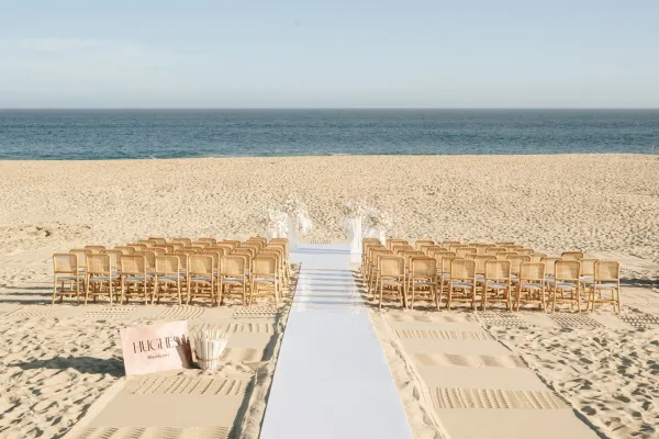 Beach ceremony setup with oceanfront wedding ceremony seating, white aisle runner, and floral pillars on sandy shore facing the horizon
