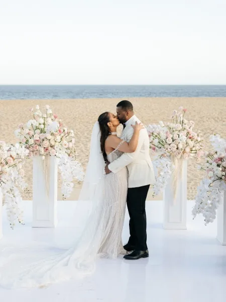 Wedding kiss portrait of bride and groom kissing under a white floral arch, her long veil flowing on an oceanfront beach platform