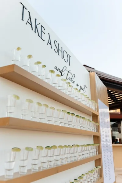 Wedding shot wall tequila shot wall with shot glasses and lime wedges on wooden shelves, plus a take a shot sign at an outdoor patio bar