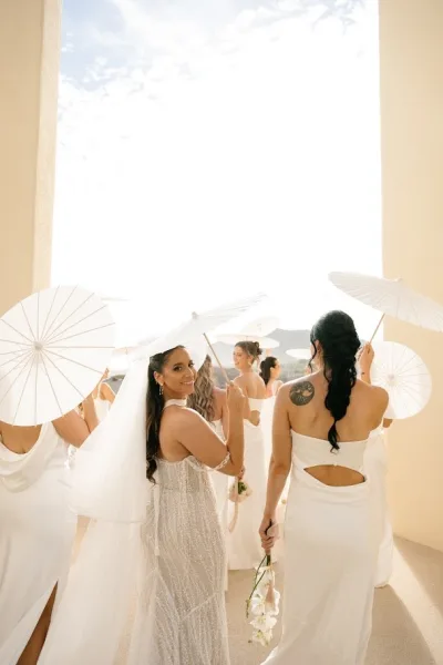 Bridesmaid portraits of women in white dresses holding parasols, walking along a beige-walled outdoor walkway under a bright sky