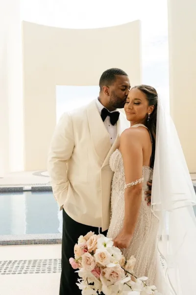 Couple portrait of groom kissing bride’s forehead as they embrace by a pool, her veil and blush rose bouquet against a stucco wall