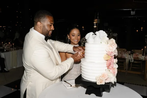 Wedding cake cutting as bride in strapless gown and groom in white tux slice a tiered cake with sugar flowers under string lights and candles