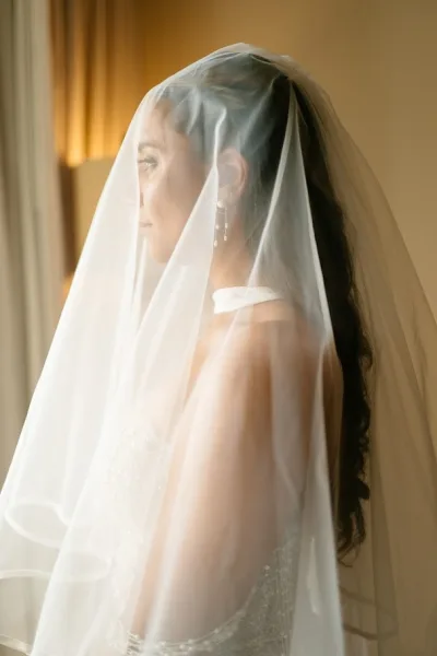 Bridal portrait of a bride wearing veil by window light, her sheer tulle veil over her face against a warm interior wall