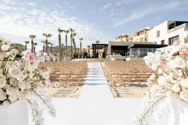 Ceremony setup for a beach wedding ceremony with a white aisle runner on sand, wooden chairs, and rose florals on pedestals under palms