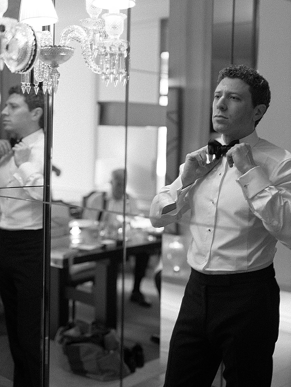 Groom getting ready adjusts his bow tie in the mirror, cufflinks on a crisp dress shirt in a softly lit hotel room with curtains