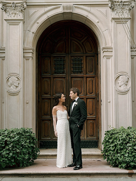 Couple portrait of bride in a strapless gown with opera gloves and groom in tuxedo, posed by arched wooden doors and stone steps
