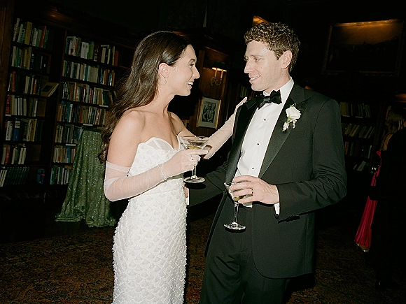 Couple portrait of newlyweds cocktail hour, bride in strapless gown and sheer gloves toasting martinis with groom in tux in a library