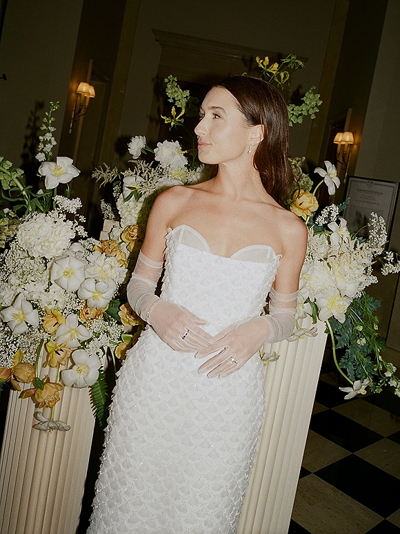 Bridal portrait in a strapless wedding dress, wearing sheer opera gloves and drop earrings, posed by a floral pedestal in an indoor venue