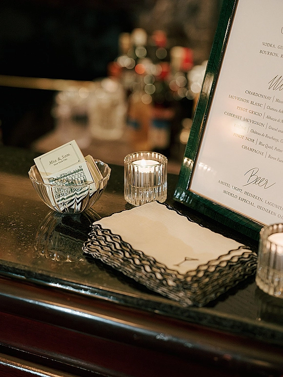 Wedding bar signage in a frame on the bar with glass votive candles, napkins, and matchbooks, liquor bottles behind in dim light