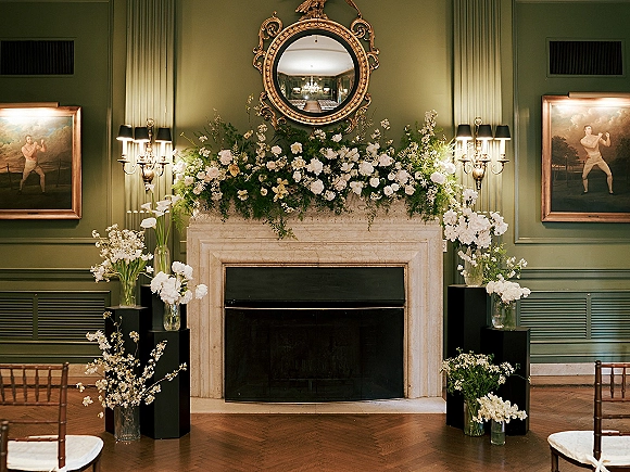 Wedding fireplace decor with fireplace wedding backdrop, white and green florals on black pedestals beneath a round gilded mirror on a green paneled wall