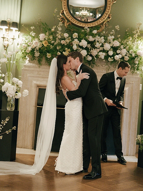 Wedding kiss as bride in veil and lace gloves kisses groom in black tuxedo by a flowered fireplace mantel with round mirror