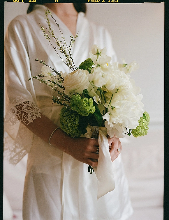 Bridal bouquet of white peonies and green viburnum tied with a white ribbon, held by a bride in a satin robe by a neutral wall