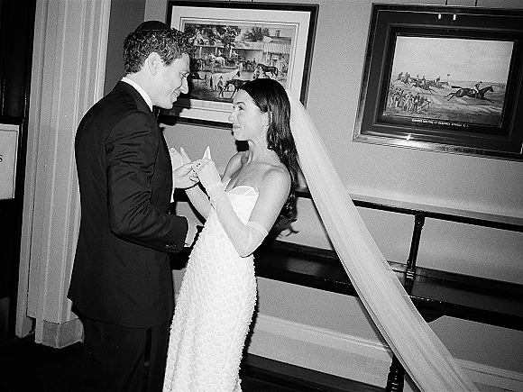 Couple portrait of bride and groom holding hands, her strapless gown with long gloves and veil, beside framed artwork indoors