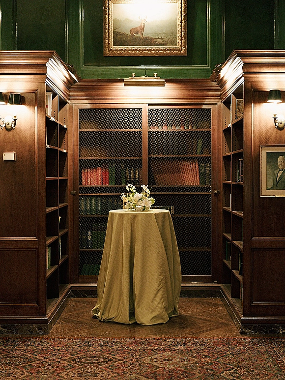 Cocktail table decor with a small floral centerpiece on a round linen-draped table in a wood-paneled library with bookshelves