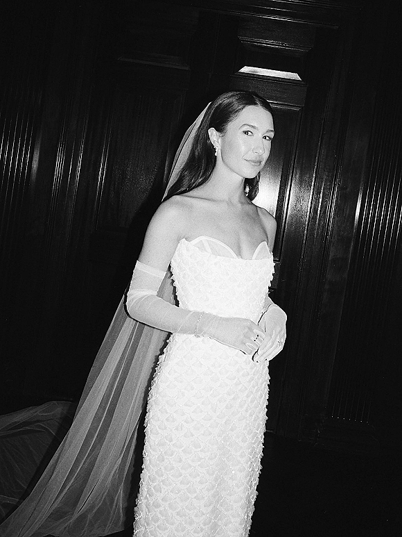 Bridal portrait in black and white of a bride looking over her shoulder in a strapless wedding dress with cathedral veil by dark wood paneling