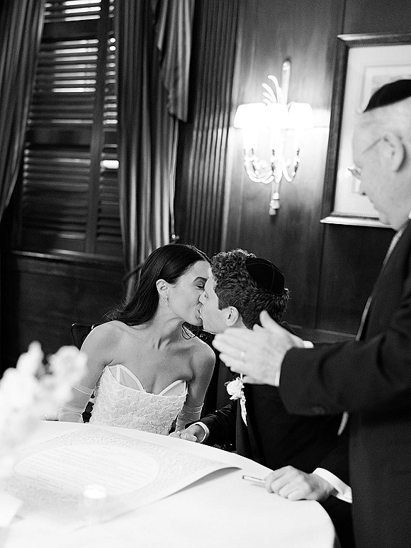Wedding kiss as bride in strapless gown and sheer opera gloves kisses groom in suit and kippah beside open license book in wood-paneled room