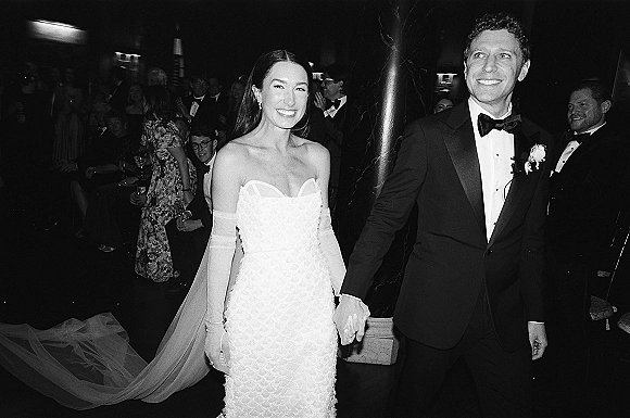 Recessional moment as bride and groom walking out, holding hands in black tie attire with long veil and opera gloves amid cheering guests indoors
