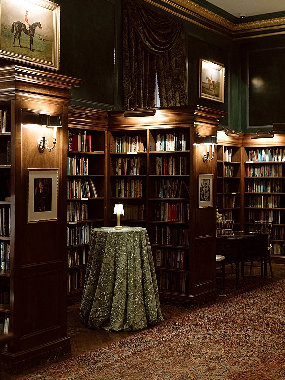 Cocktail hour decor with a round cocktail table in a patterned tablecloth, lamp centerpiece, and shelves in a dark wood library room