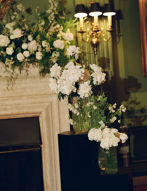 Wedding floral arrangements of white wedding flowers and greenery in glass vases on pedestals by a fireplace mantel with framed art