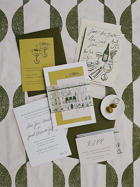 Invitation suite flat lay with envelopes, RSVP and save the date cards, and a ring on green leaf print fabric, beside a plate of olives