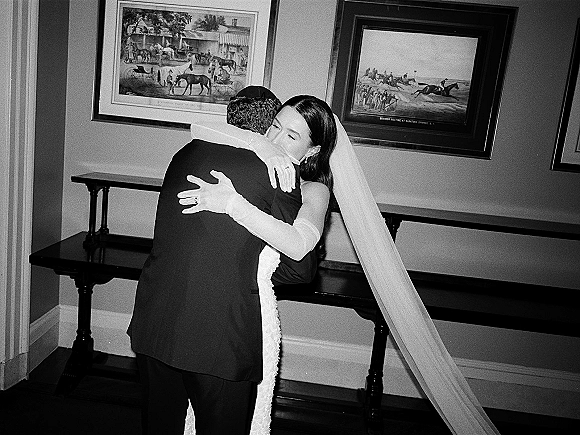 Wedding hug as bride in veil and long gloves embraces groom in black tuxedo and kippah indoors by framed artwork and molding