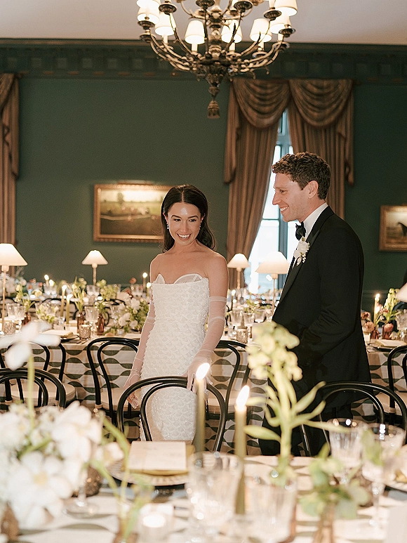 Bride and groom at wedding reception couple’s candlelit table, bride in strapless gown and sheer opera gloves under chandelier in green dining room