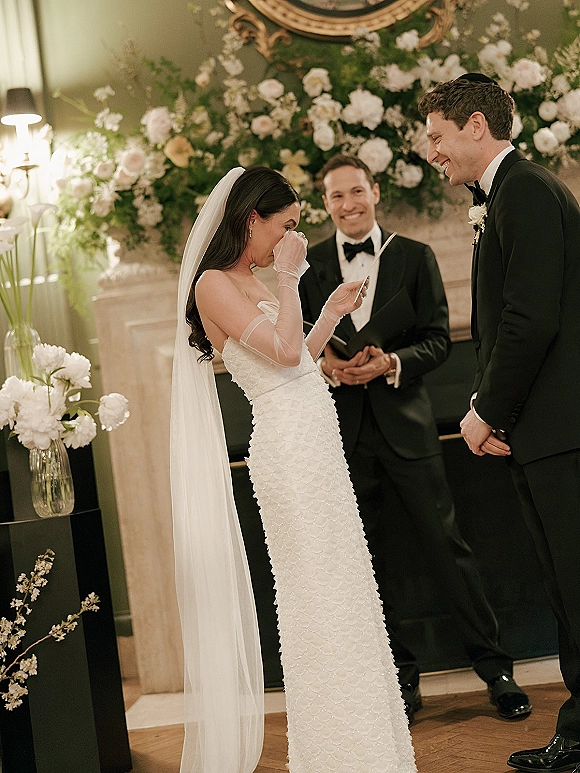 Wedding vows as bride reads from a vow card, wiping tears, while groom smiles beside her in front of a fireplace mantel garland