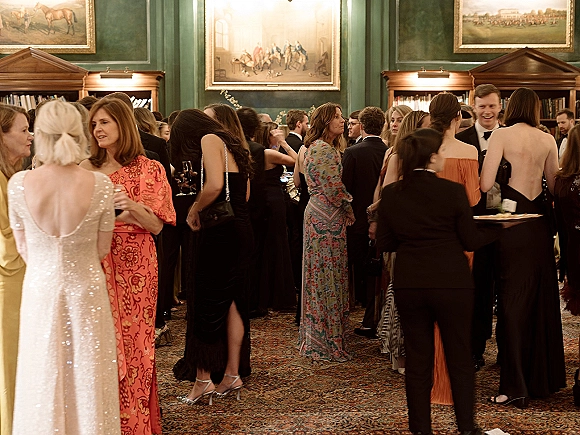 Wedding cocktail hour with wedding guests mingling in tuxedos and formal gowns, holding cocktail glasses in a cozy library room with bookshelves