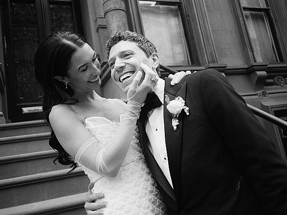 Couple portrait in a black and white wedding portrait style, bride laughing as she touches groom’s face on stone steps by windows