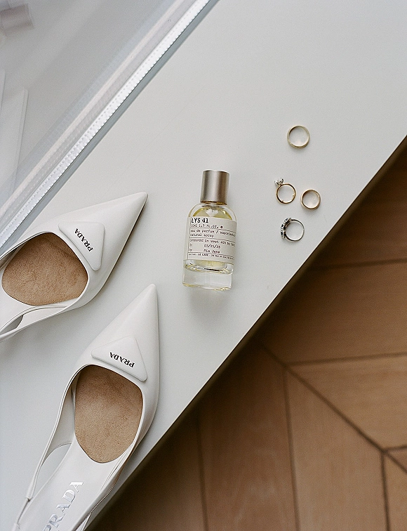 Wedding rings flatlay featuring a wedding ring detail photo with engagement ring beside perfume bottle and white pointed-toe heels in window light