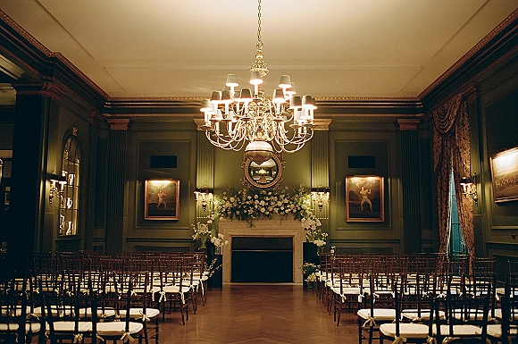 Indoor ceremony setup with wedding ceremony chairs in Chiavari rows facing a fireplace floral arrangement beneath a chandelier in a green-paneled room