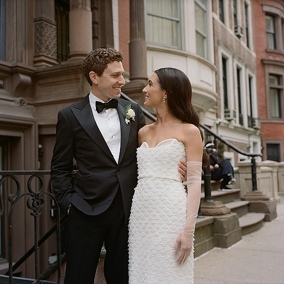Couple portrait of bride and groom portrait on a brownstone stoop, embracing by iron railing; groom in black tuxedo with boutonniere