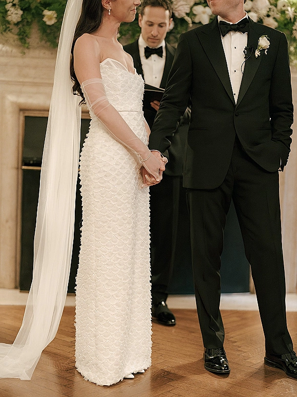 Wedding vows as bride and groom hold hands during an indoor wedding ceremony, officiant before a fireplace mantel and greenery backdrop