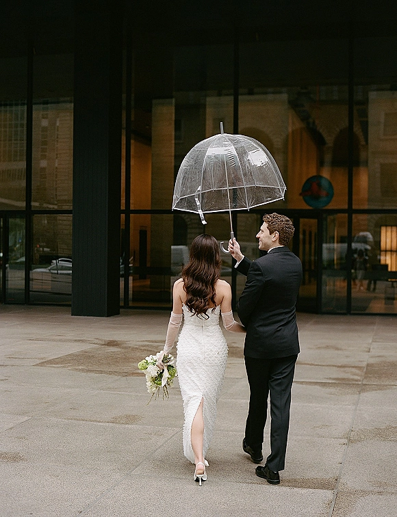 Couple portrait of bride and groom walking away under a clear umbrella, bride holding bouquet on wet city sidewalk by glass building