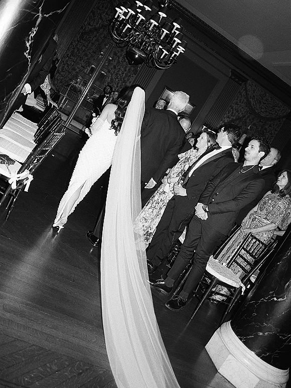 Wedding processional with bride walking down aisle in a strapless gown and cathedral veil, holding bouquet as guests stand under chandelier