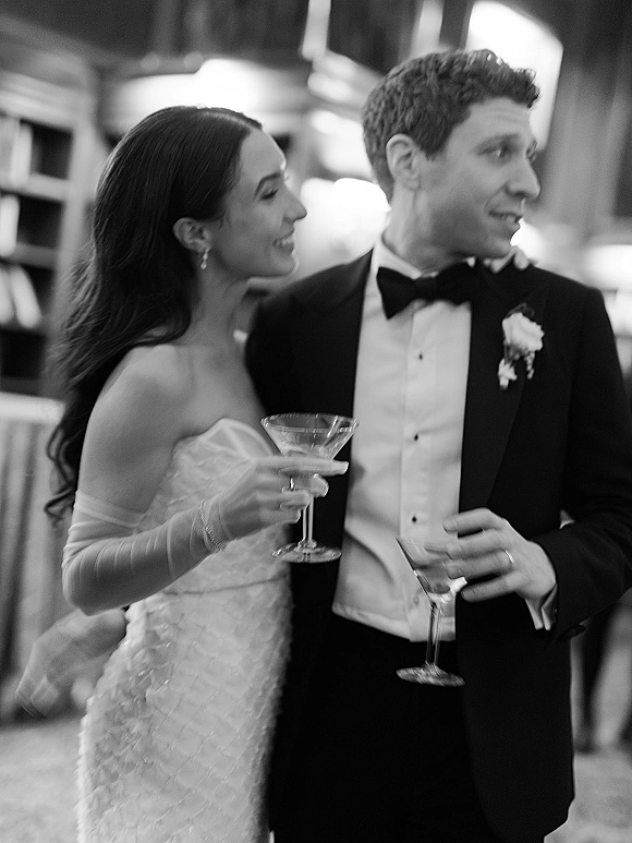 Wedding couple portrait of bride and groom with drinks, laughing in a black-tie indoor reception with blurred guests behind them