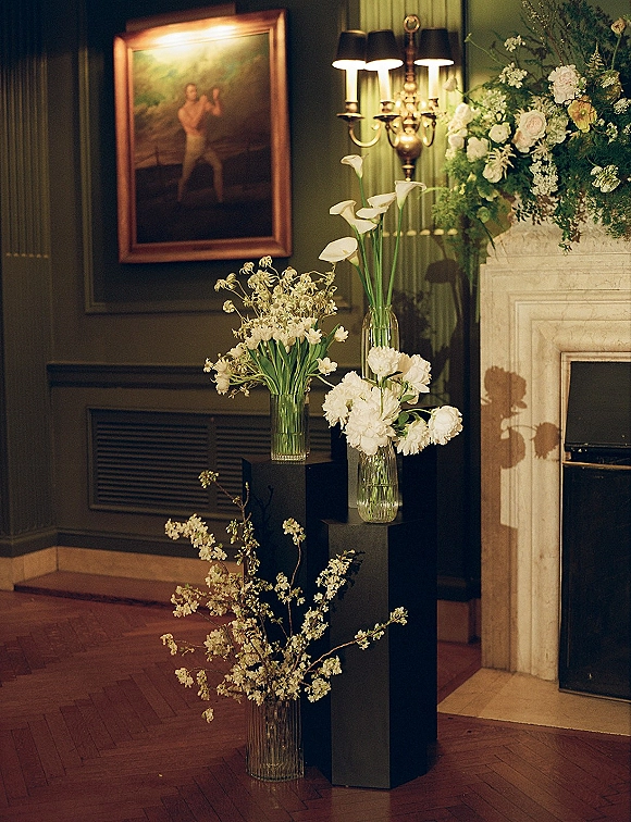 Wedding floral arrangement with white wedding flowers and calla lilies on black plinths by a marble fireplace against dark paneled walls