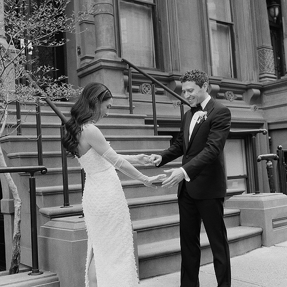 Wedding first look as groom adjusts bride’s opera glove on stone townhouse steps, both holding hands in tuxedo and strapless lace dress
