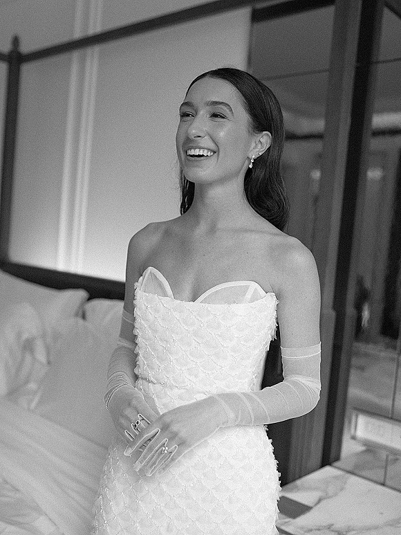 Bridal portrait in black and white of a laughing bride in a strapless textured gown with sheer opera gloves beside a window-lit bed