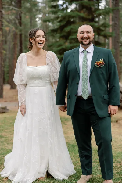 Couple portrait of bride and groom holding hands and laughing, her puff-sleeve embroidered gown and his suit on a forest lawn