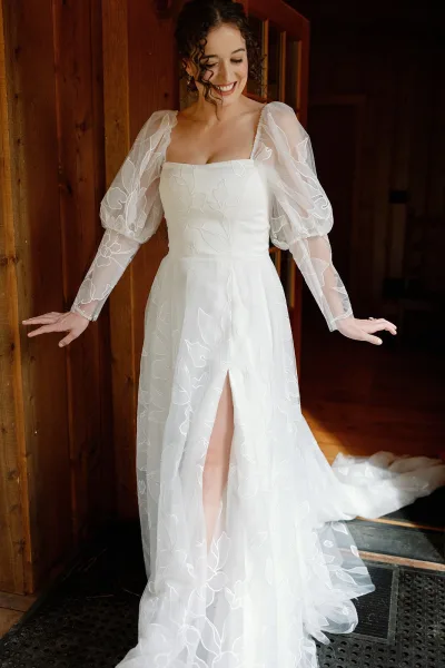 Bridal portrait of a smiling bride in doorway wearing a sheer puff-sleeve wedding dress with embroidered tulle overlay by a wood-paneled hall