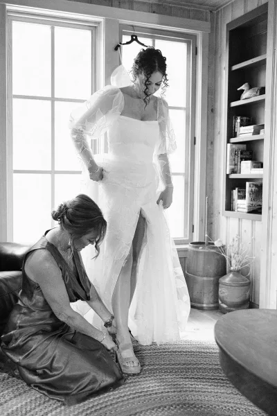 Bridal getting ready as a bridesmaid helps the bride put on shoes, adjusting her puff-sleeve wedding dress in soft window light living room