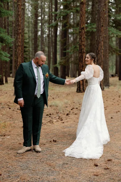 Couple portrait of bride and groom holding hands, bride laughing in puff sleeve dress and gloves beside dark green suit in pine forest