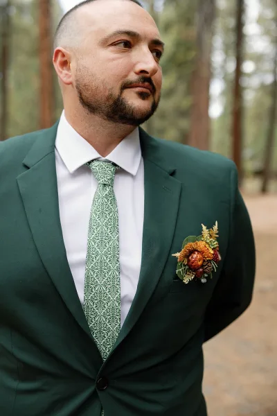 Groom portrait in a green wedding suit with patterned tie and boutonniere, standing on an outdoor path framed by forest trees