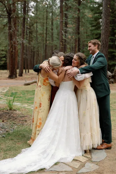Wedding group hug as the bride hugs family, dress train spread out, with floral bridesmaid dresses and groom in pine forest on a stone path