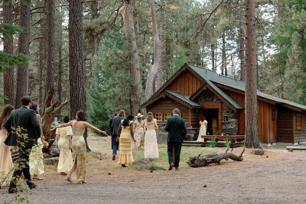 Wedding guests arriving along a dirt path in a pine forest, dressed in suits and pastel dresses near a rustic cabin with string lights